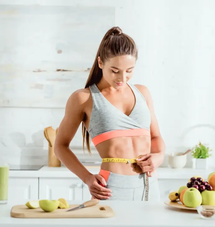 A woman measuring her waist beside a counter of fresh fruit and vegetables
